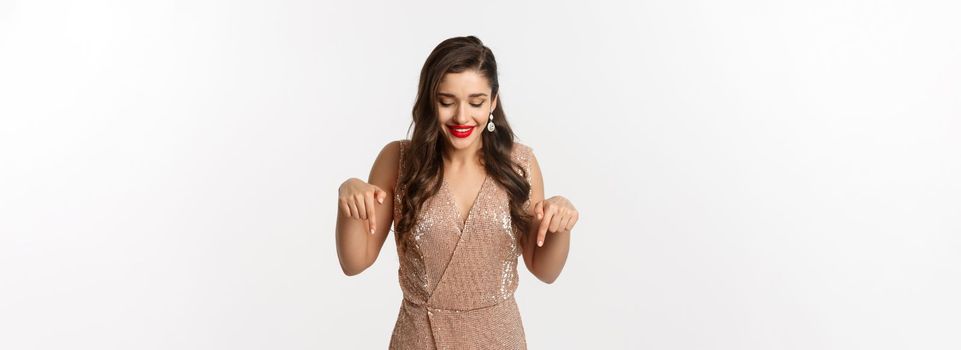 Beautiful young woman with red lipstick, wearing elegant dress for christmas party, pointing fingers down and smiling pleased, standing over white background.