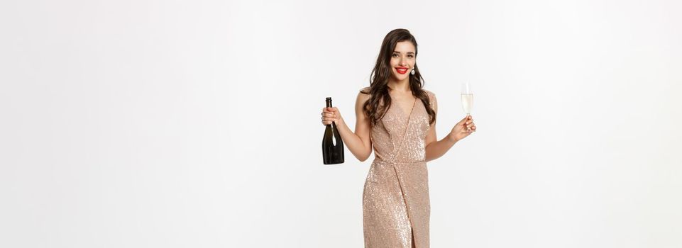 Christmas party and celebration concept. Full length of elegant woman with red lips, luxury dress, holding glass of champagne and bottle, white background.