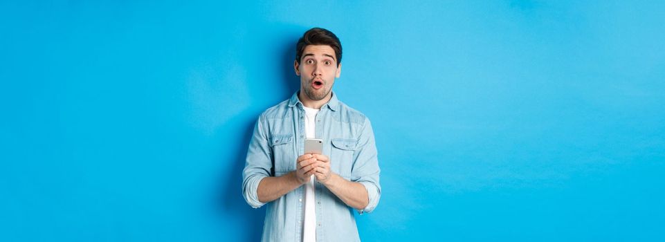 Surprised man in casual clothes looking astounded, holding smartphone, standing against blue background.