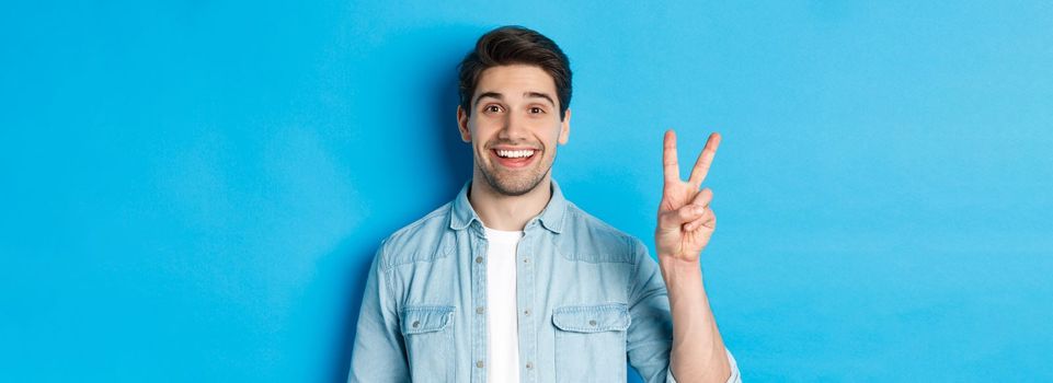 Close-up of handsome man smiling, showing fingers number two, standing over blue background.