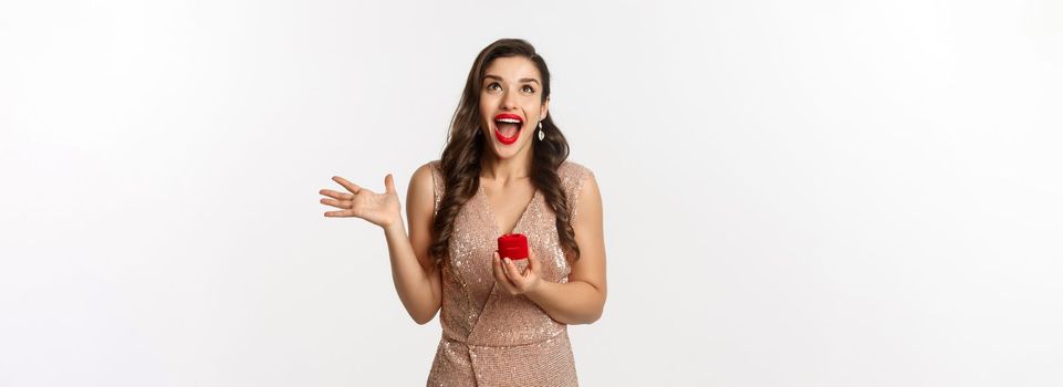 Image of excited woman receiving engagement ring and marriage proposal, scream of joy and happiness, wearing evening dress and red lipstick, standing over white background.