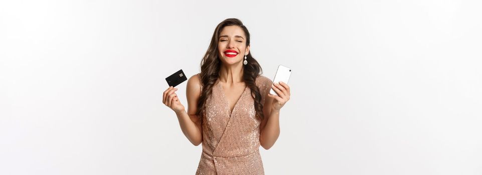 Online shopping and holidays concept. Satisfied and happy woman in elegant dress smiling, using credit card and mobile phone, standing over white background.