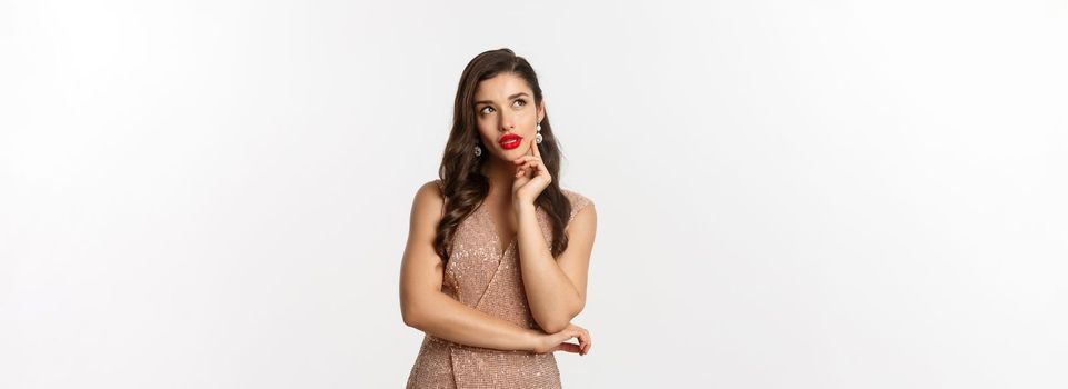 Christmas, holidays and celebration concept. Young woman looking thoughtful at upper left corner, wearing evening dress for party, thinking about New Year eve, white background.