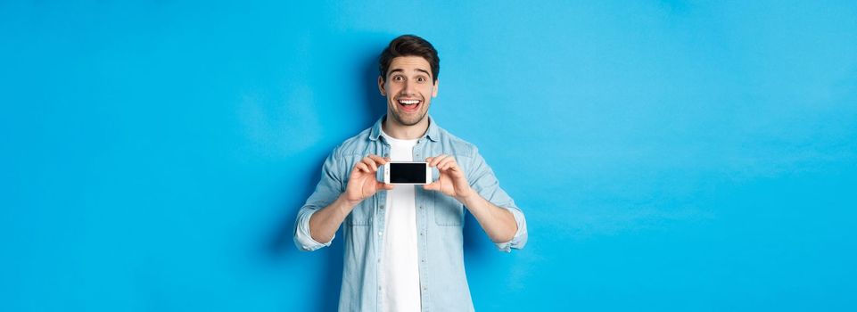 Amazed smiling man showing smartphone screen, internet promo offer, standing against blue background.