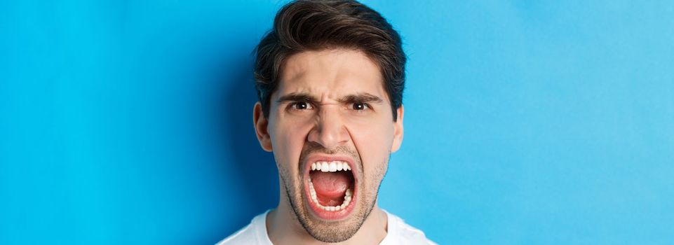 Head shot of angry man shouting and looking with hatred, standing mad against blue background.