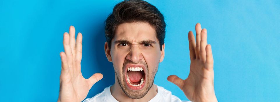 Headshot of man showing anger and frustration, yelling with outraged face, standing over blue background.