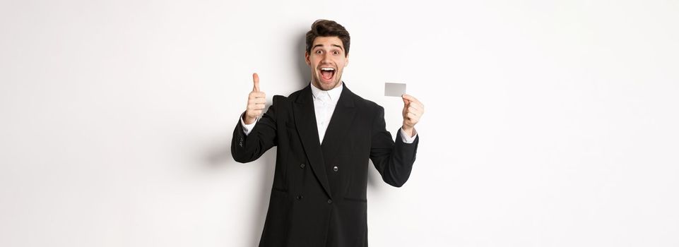 Image of good-looking businessman in black suit, showing thumbs-up and credit card, recommending bank, standing against white background.