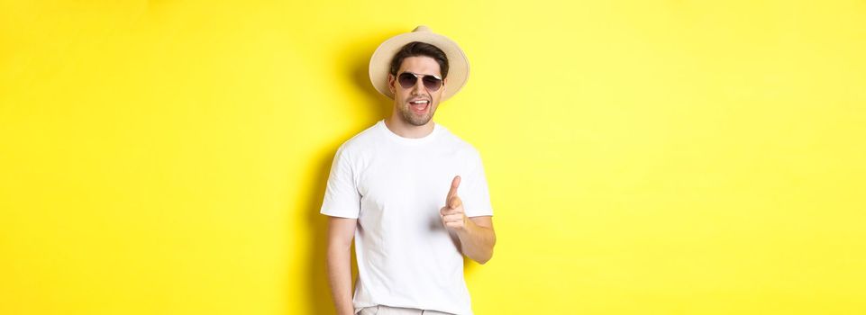 Confident and cheeky guy on vacation flirting with you, pointing finger at camera and winking, wearing summer hat with sunglasses, yellow background.