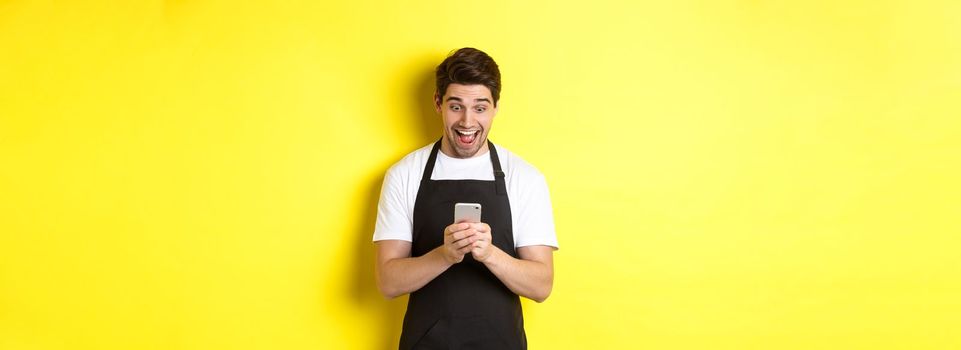 Barista looking surprised as reading message on mobile phone, standing in black apron against yellow background.