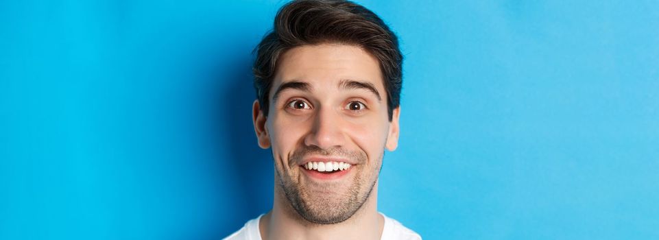 Head shot of handsome male model looking amazed, smiling wondered, standing over blue background. Copy space
