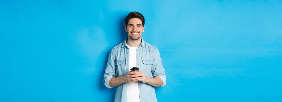 Young happy man drinking coffee from cafe takeaway, smiling pleased, standing against blue background.