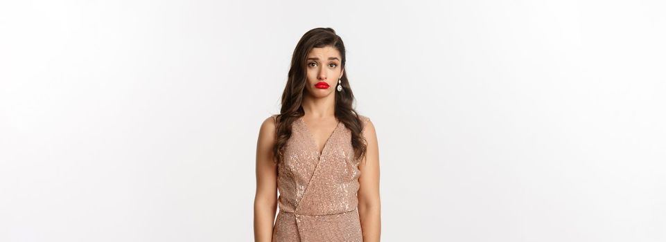 Celebration and party concept. Sad and confused young woman in glamour dress staring at camera distressed, standing against white background.