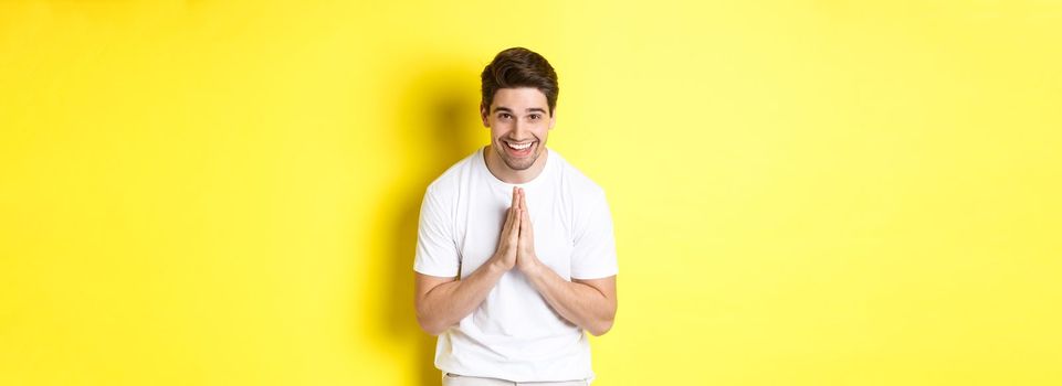Handsome guy saying thank you, bowing and holding hands in namaste gesture, express gratitude, standing over yellow background.