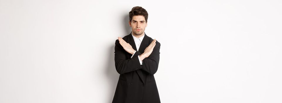 Portrait of serious and concerned man in black suit, showing stop gesture and frowning, making cross to prohibit or decline something, standing over white background.