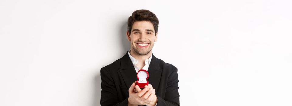 Image of handsome man looking romantic, open small box with engagement ring, making a proposal and smiling, standing against white background.