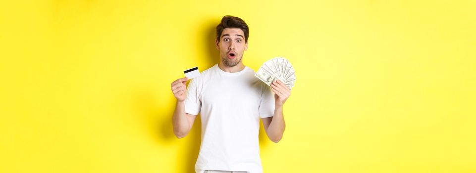 Amazed man holding credit card and money, looking surprised, standing over yellow background.