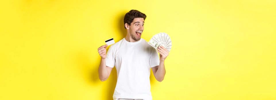 Cheerful guy looking at money, holding credit card, concept of bank credit and loans, standing over yellow background.