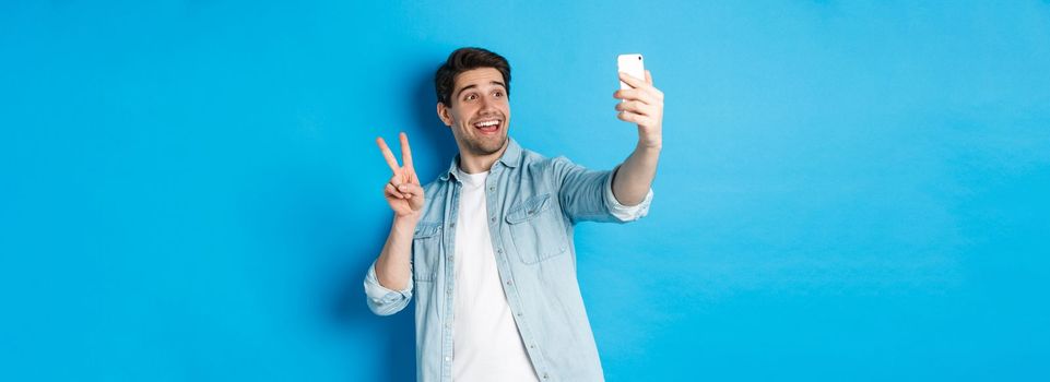 Happy man taking selfie and showing peace sign on blue background, holding mobile phone.