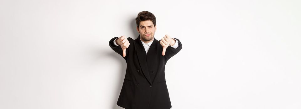 Portrait of skeptical and disappointed man in black suit, frowning upset, showing thumbs-down, dislike something bad, standing over white background.