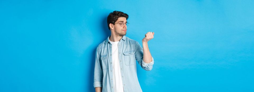 Young handsome man in glasses looking at his fingernails, checking manicure, standing over blue background.