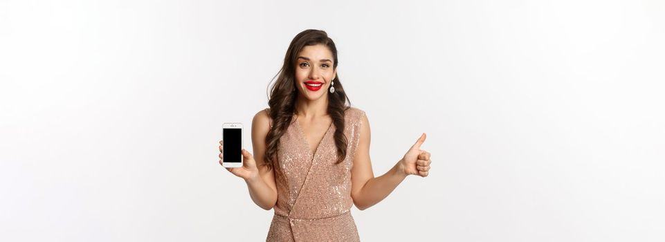 Online shopping. Beautiful young woman in party dress and makeup, showing thumbs up and mobile screen, praising good app, white background.