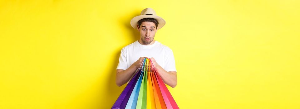 Happy man looking surprised at shopping bags, buying souvenirs on vacation, standing over yellow background.