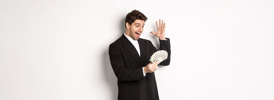 Image of successful businessman in black suit, looking at money and triumphing, earn cash, standing over white background.