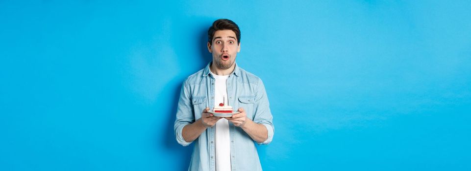 Man holding birthday cake and looking surprised, making a wish on lit candle, standing over blue background.