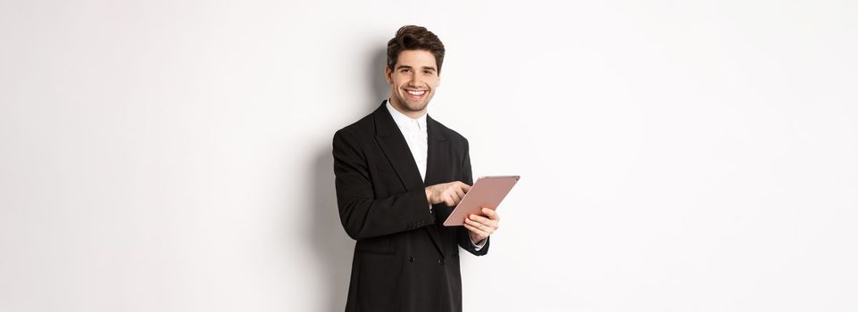 Image of handsome businessman in black suit, using digital tablet and smiling, standing against white background.