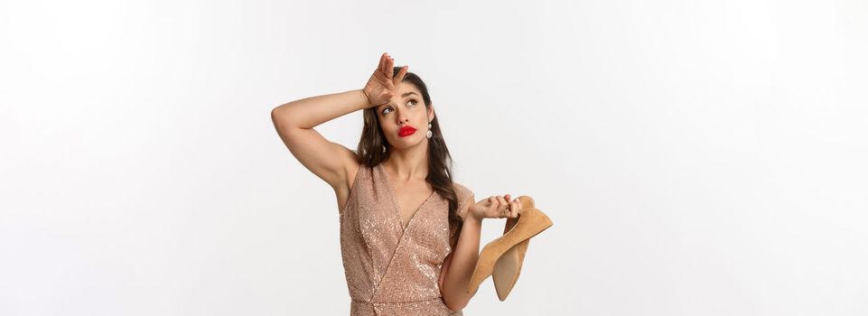 Party and celebration concept. Tired woman in elegant dress take off pair of heels, holding hand on forehead, express fatigue, standing over white background.