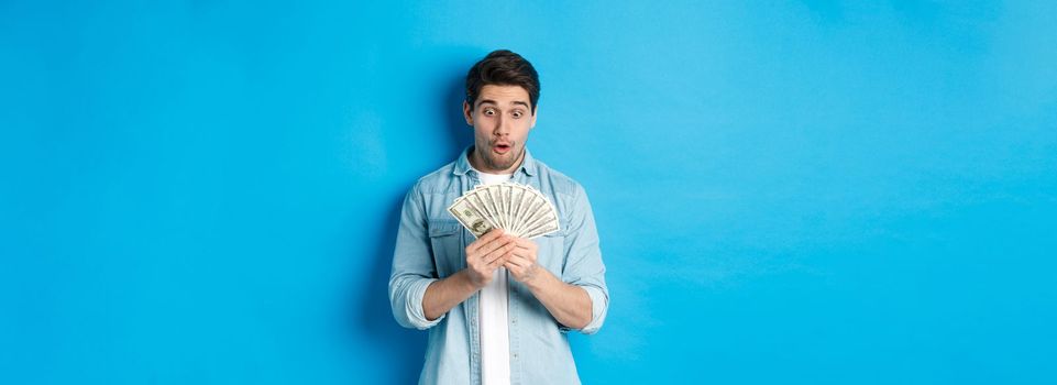 Excited successful man counting money, looking satisfied at cash and smiling, standing over blue background.