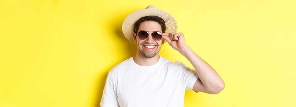 Concept of tourism and vacation. Close-up of handsome man tourist looking happy, wearing sunglasses and summer hat, standing over yellow background.