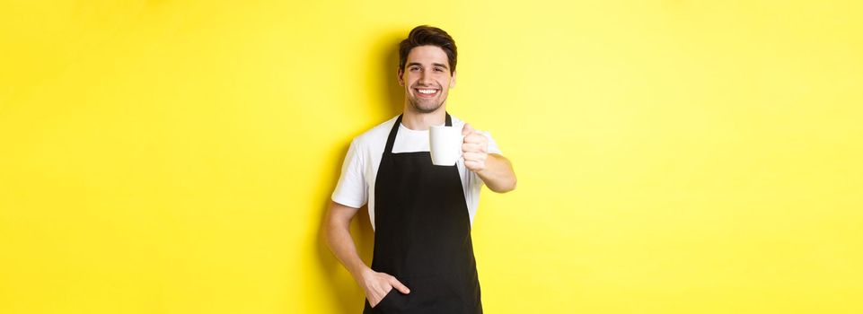 Handsome barista in black apron giving you cup of coffee and smiling, standing over yellow background.