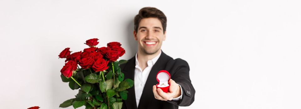 Close-up of attractive man in suit, holding bouquet of roses giving an engagement ring, proposing to girlfriend, standing against white background.