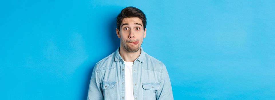 Close-up of young man making funny expressions, showing tongue and looking at camera, standing over blue background.