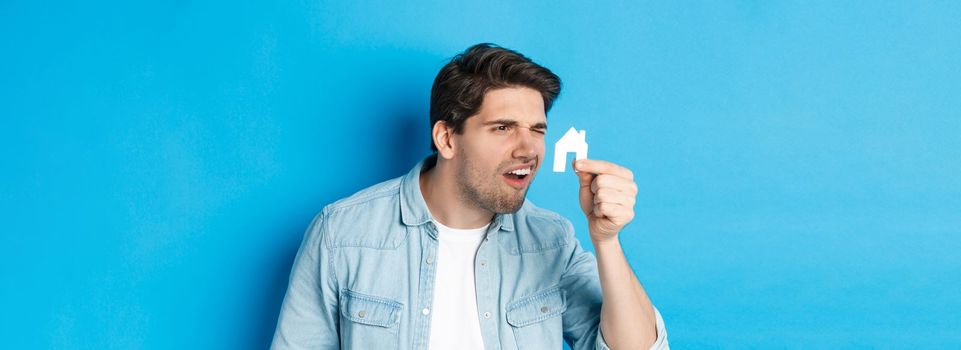 Real estate concept. Man looking at tiny house maket with confused face, standing puzzled against blue background.