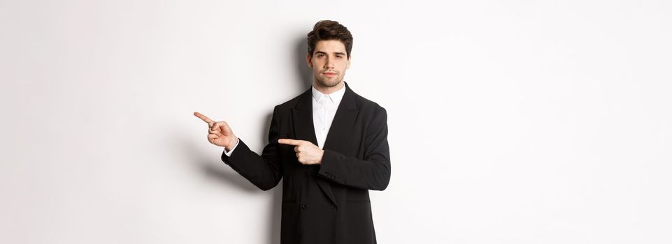 Image of handsome businessman in black suit, pointing fingers right and looking at camera, standing against white background.