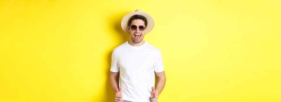 Confident and cheeky guy on vacation flirting with you, pointing finger at camera and winking, wearing summer hat with sunglasses, yellow background.