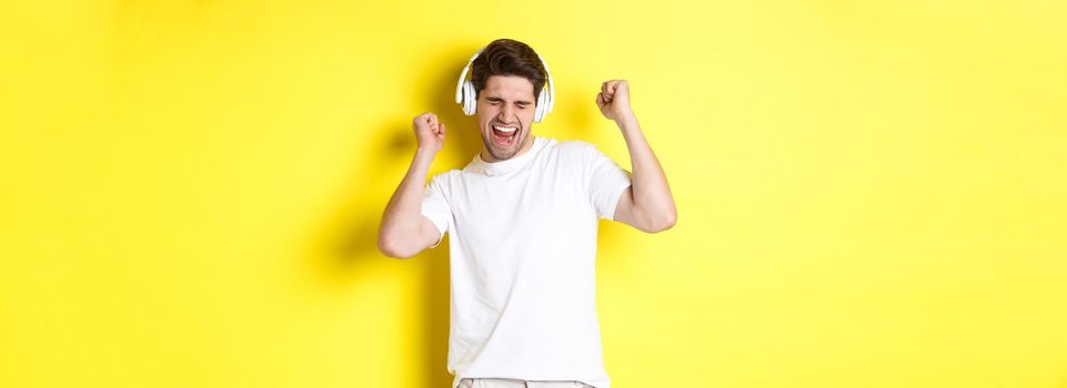 Happy man dancing and listening music in white headphones, standing over yellow background. Copy space