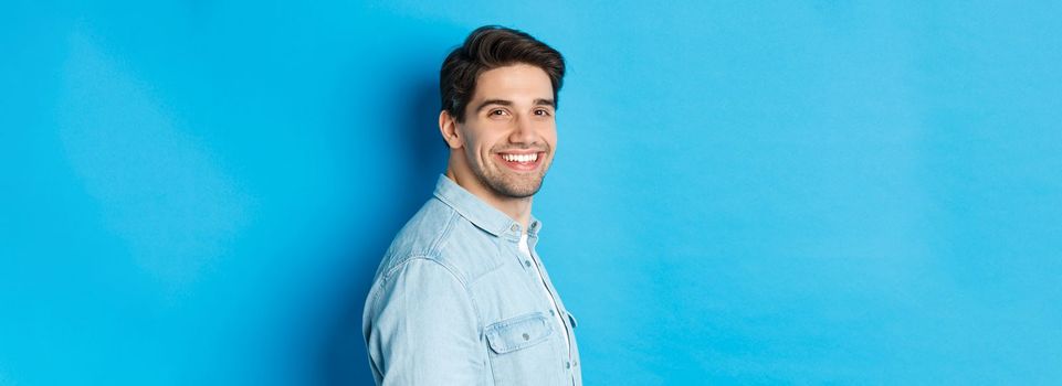 Handsome and confident guy turn head at camera, smiling happy, standing over blue background.