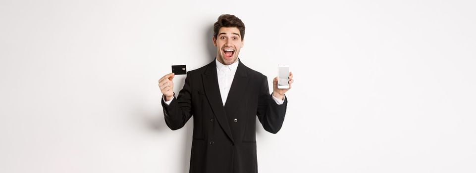 Image of handsome businessman in black suit, looking excited and showing credit card with mobile phone screen, standing against white background.