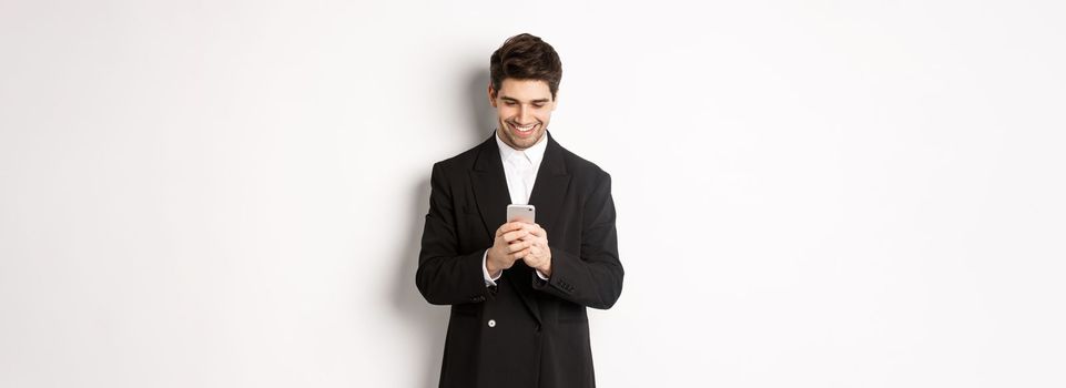Portrait of handsome stylish businessman in black suit, writing a message, smiling and looking at smartphone, standing over white background.