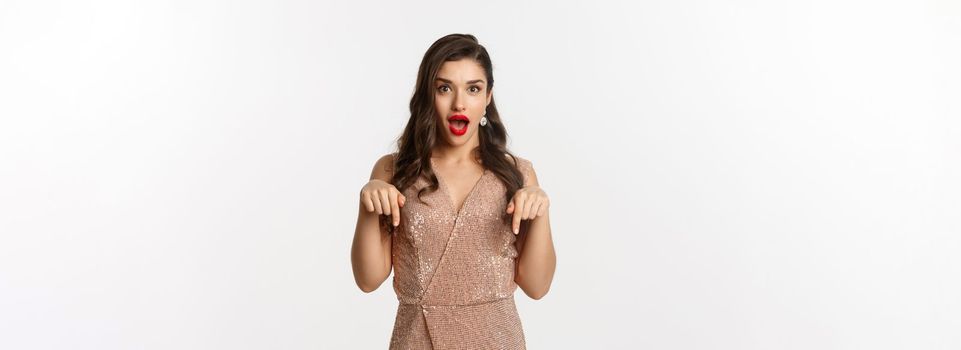 Amazed beautiful woman in glamour dress celebrating christmas party, pointing fingers down at promo, standing over white background.