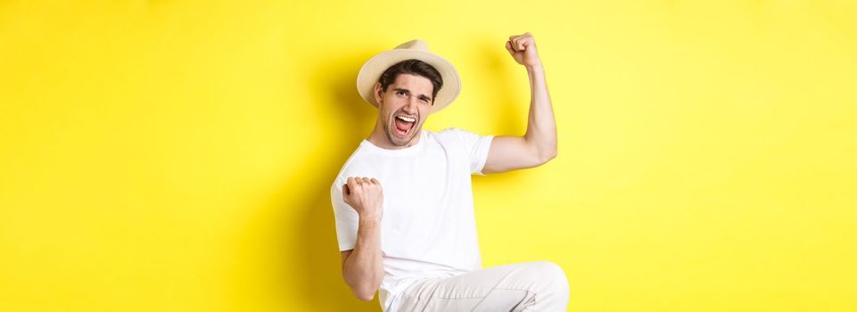 Concept of tourism and vacation. Lucky male tourist winning trip, rejoicing and saying yes, making fist pump while triumphing, standing against yellow background.