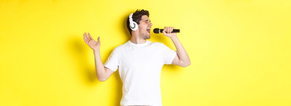 Passionate guy in headphones holding microphone, singing karaoke song, standing over yellow background in white clothes.
