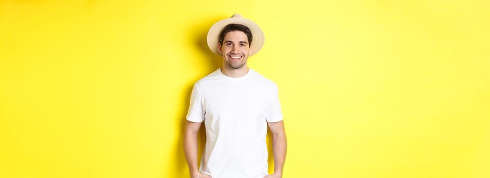 Young handsome guy tourist looking happy, wearing straw hat for travelling, standing against yellow background. Copy space