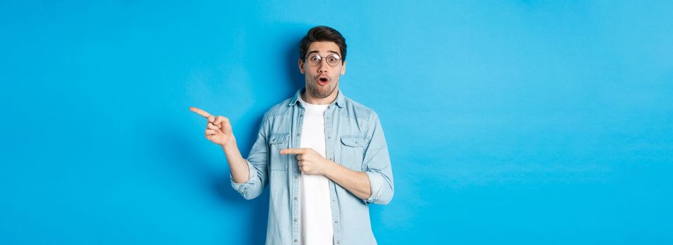 Surprised man in glasses pointing right at copy space, showing promo offer on blue background, standing over blue background.