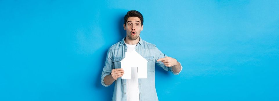 Insurance, mortgage and real estate concept. Surprised young man pointing at house card model and looking at camera, standing against blue background.