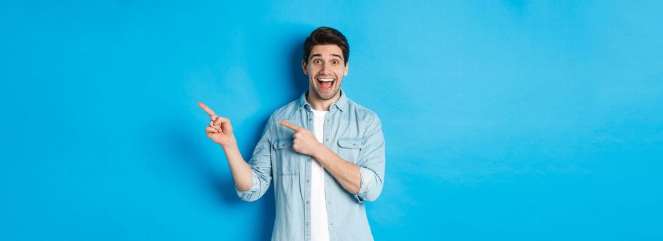 Image of excited handsome man in casual outfit, showing advertisement, pointing fingers right at copy space and smiling, standing against blue background.