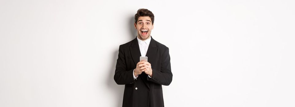 Image of excited handsome guy receiving promo offer on mobile phone, looking amazed and smiling, standing against white background.
